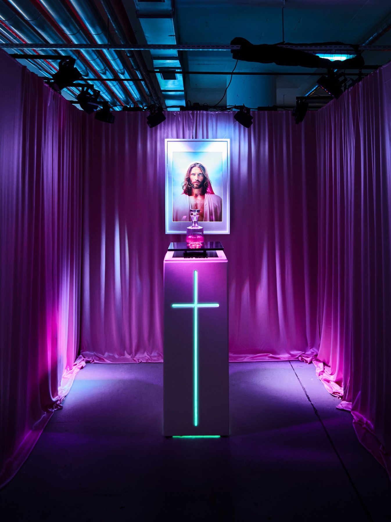 ai_am_jesus installation: portrait in a glowing frame above a decanter on a plinth with a neon cross, framed by pink curtains