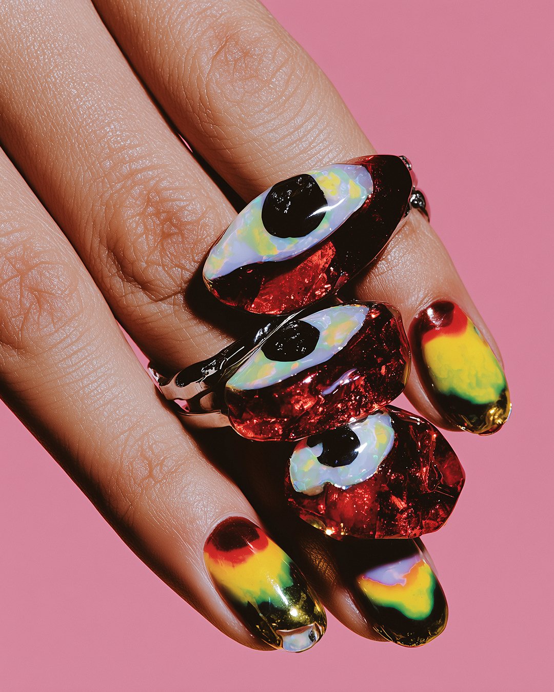 Hand with three glossy red eye-shaped rings and iridescent nails against a pink background